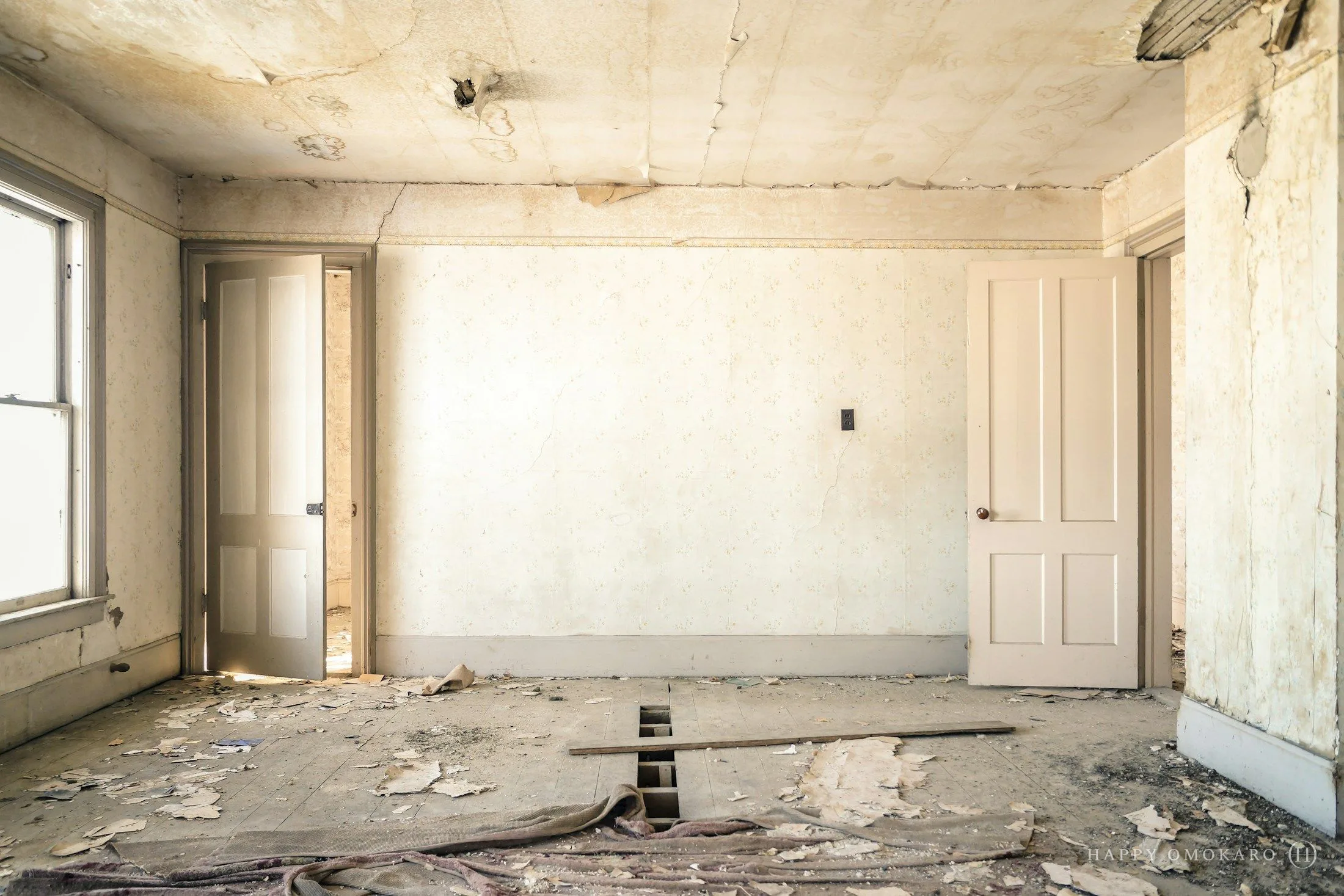 An abandoned room with peeling wallpaper, two open doors and a worn wooden floor