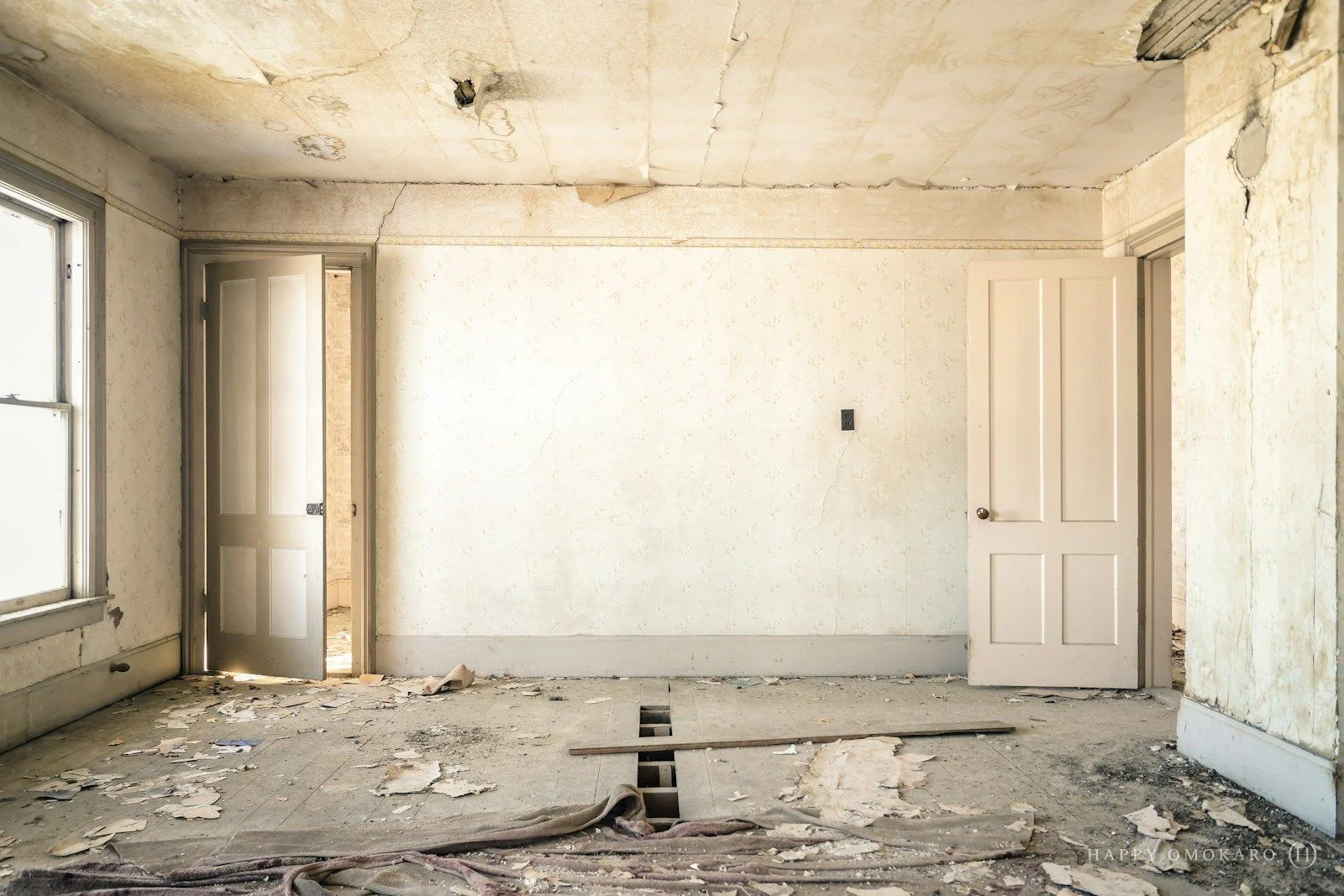 An abandoned room with peeling wallpaper, two open doors and a worn wooden floor